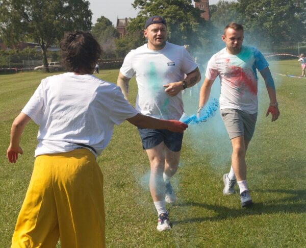 two men run towards camera, their clothes are covered in multi paint powder marks. A woman throws blue paint powder at them