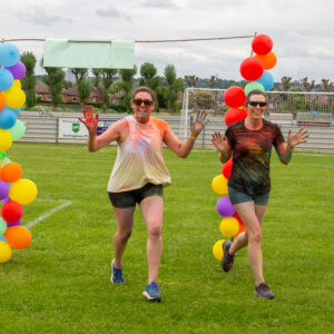 2 women running through a balloon arch. Their clothes are covered in paint powder.