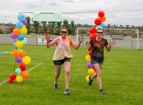 2 women running through a balloon arch. Their clothes are covered in paint powder.
